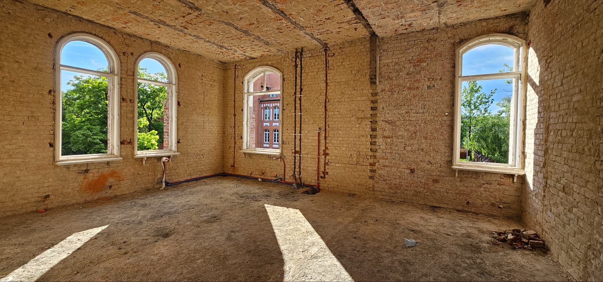 Wnętrze w trakcie remontu: surowe ceglane ściany, trzy łukowe okna z widokiem na zieleń, instalacje rurowe. Podłoże ziemne, naturalne światło słoneczne.