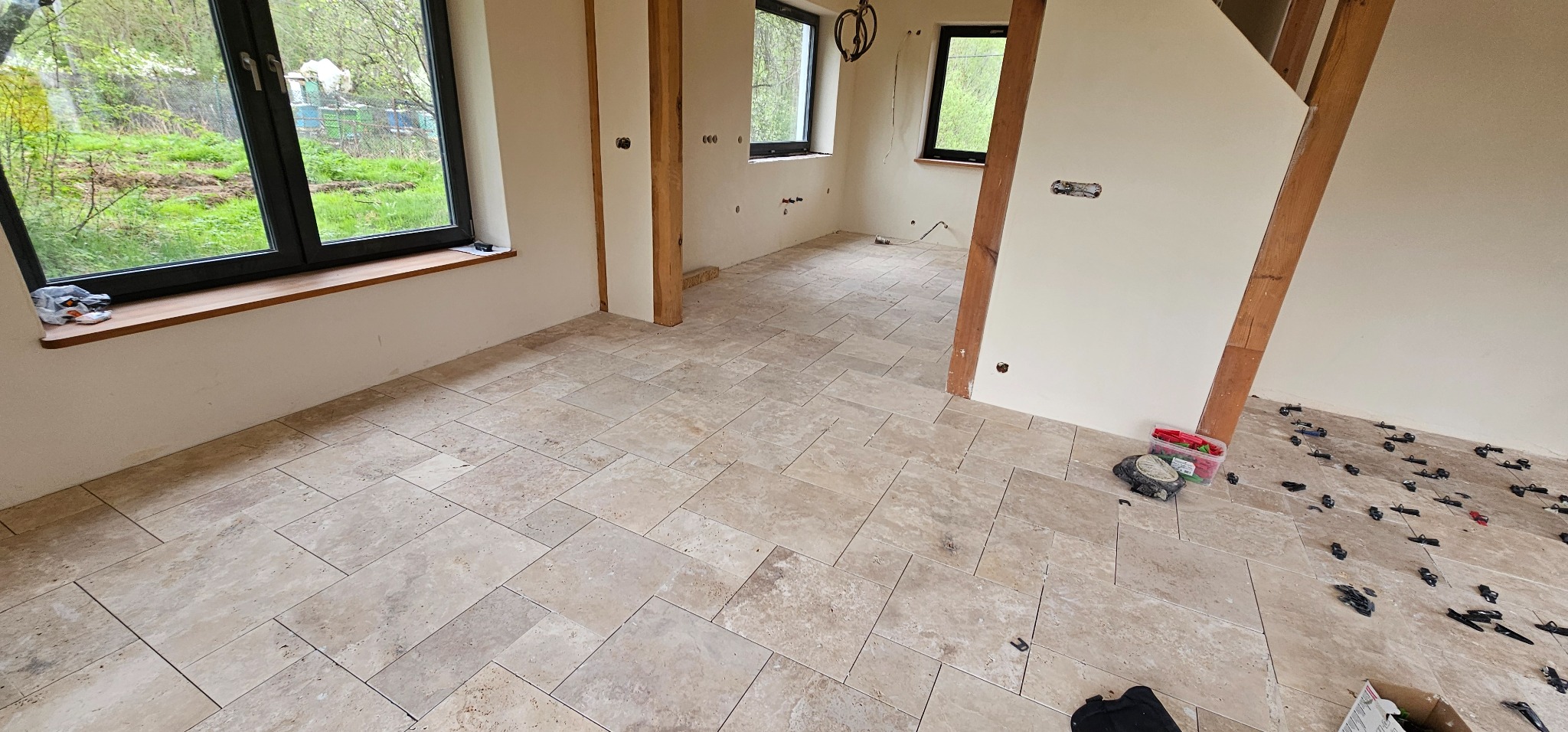 Beżowe płytki podłogowe ułożone w jodełkę w jasnym pokoju z czarnymi oknami. Widoczne narzędzia glazurnicze i elementy drewniane konstrukcji domu.