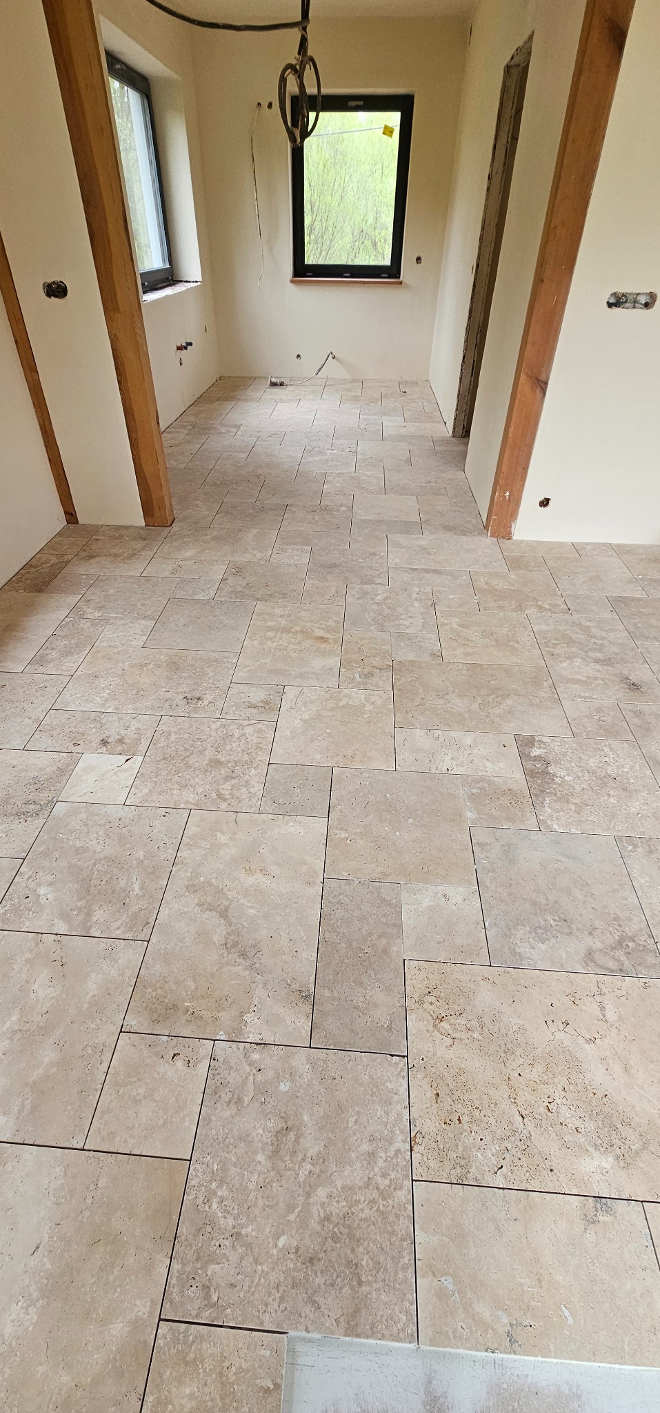 Ułożona podłoga z trawertynu w jasnym pomieszczeniu z oknami i drewnianymi elementami konstrukcyjnymi. Widoczne przygotowania do dalszych prac wykończeniowych.