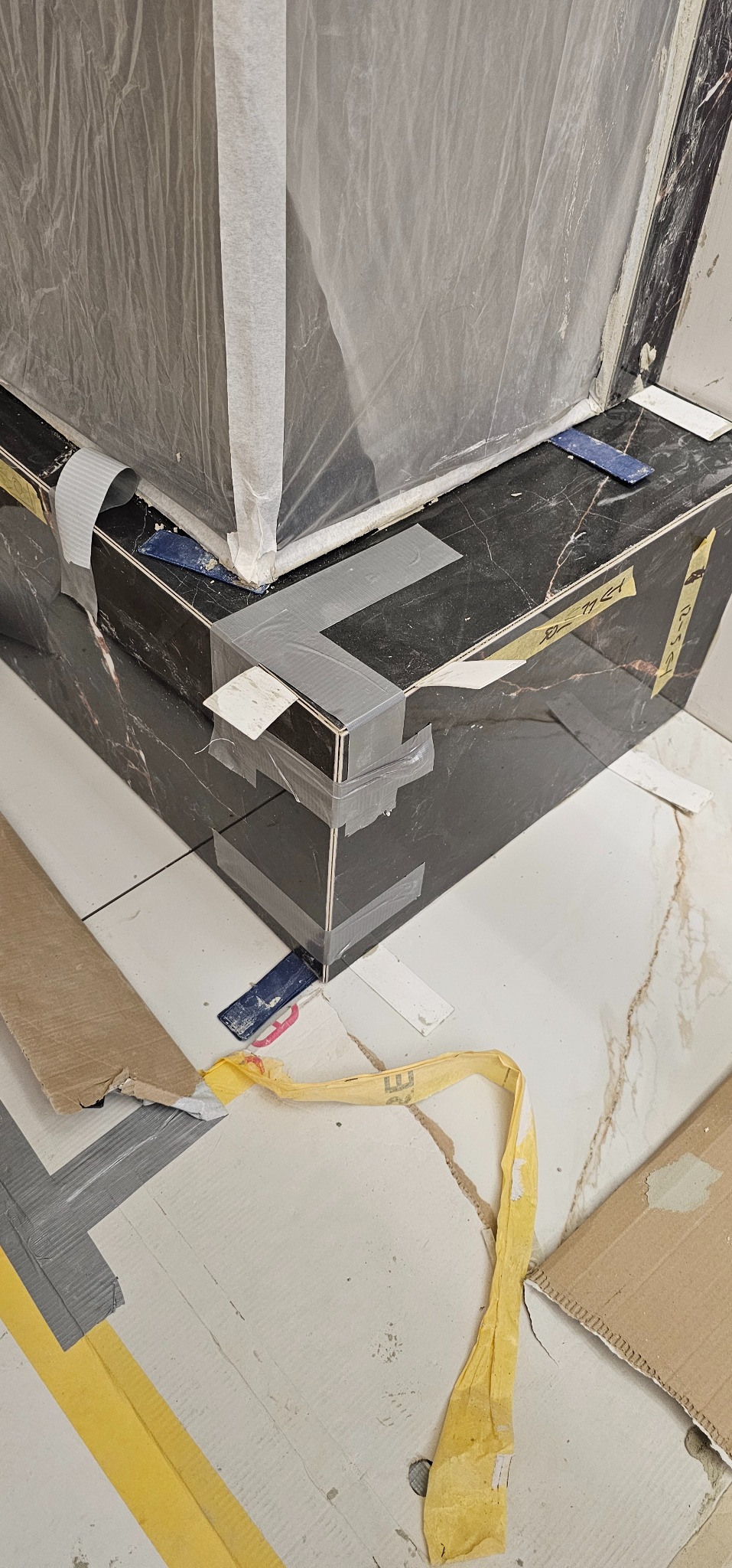 Fragment łazienki w trakcie wykończenia: czarne płytki imitujące marmur, zabezpieczone folią i taśmą, białe płytki podłogowe, resztki materiałów budowlanych.