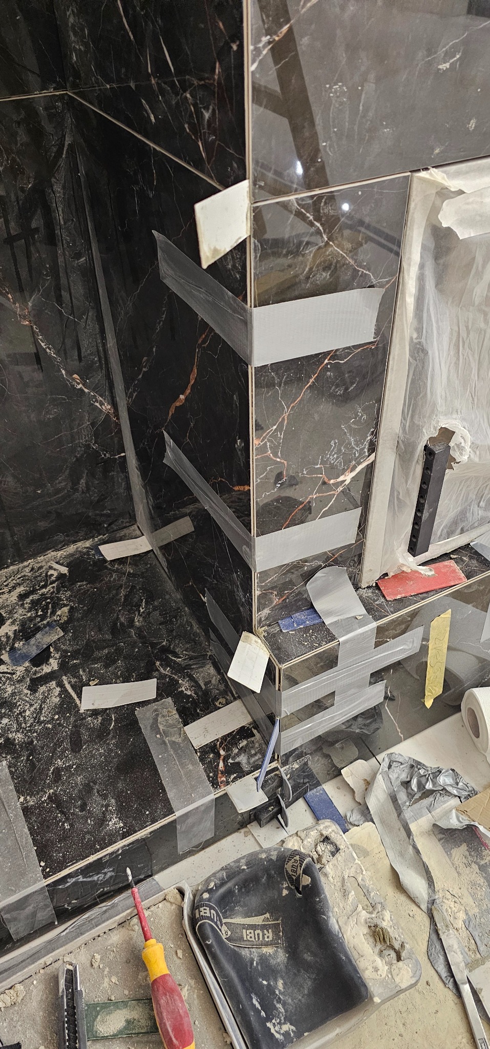 Proces układania czarnych, marmurkowych płytek w rogu pomieszczenia, zabezpieczonych taśmą. Widoczne narzędzia glazurnicze i akcesoria, co wskazuje na trwające prace wykończeniowe.