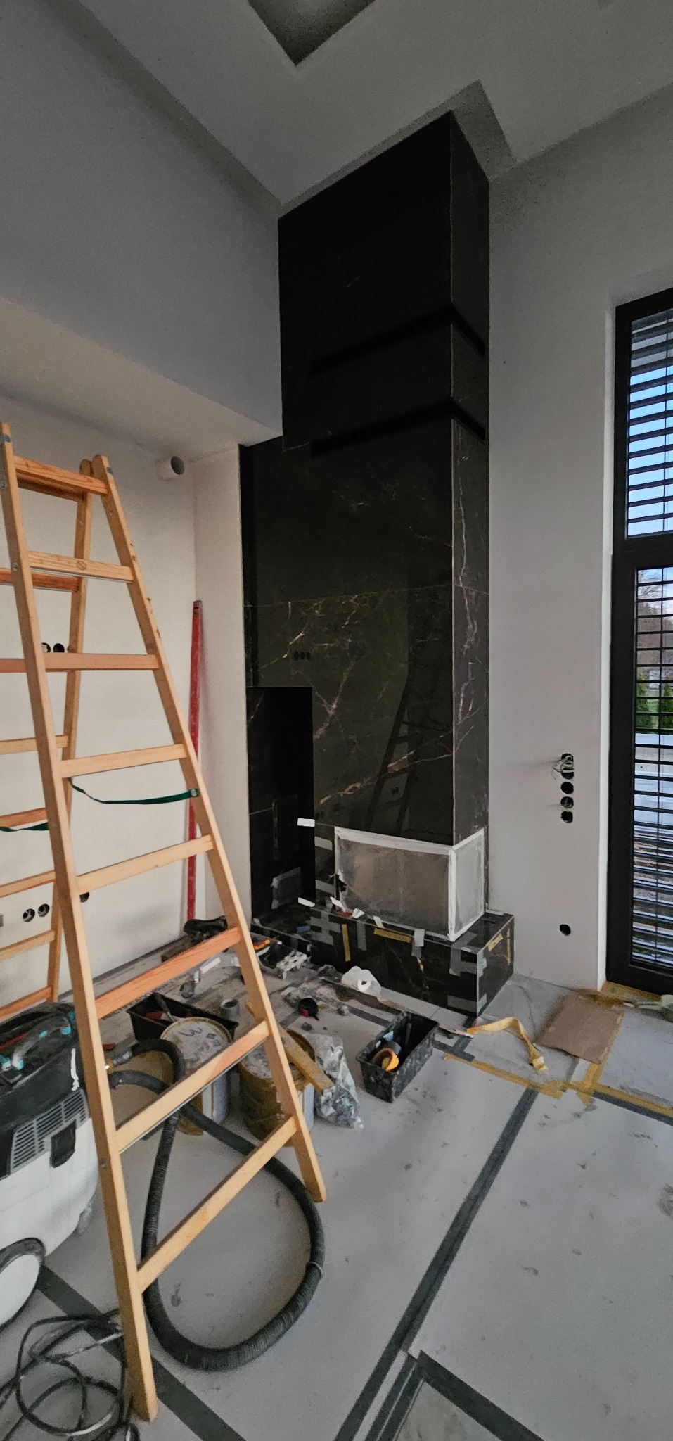 Wnętrze w trakcie wykończenia: obłożony czarnym marmurem kominek z widoczną konstrukcją wkładu, drabina malarska, narzędzia i materiały budowlane na podłodze.