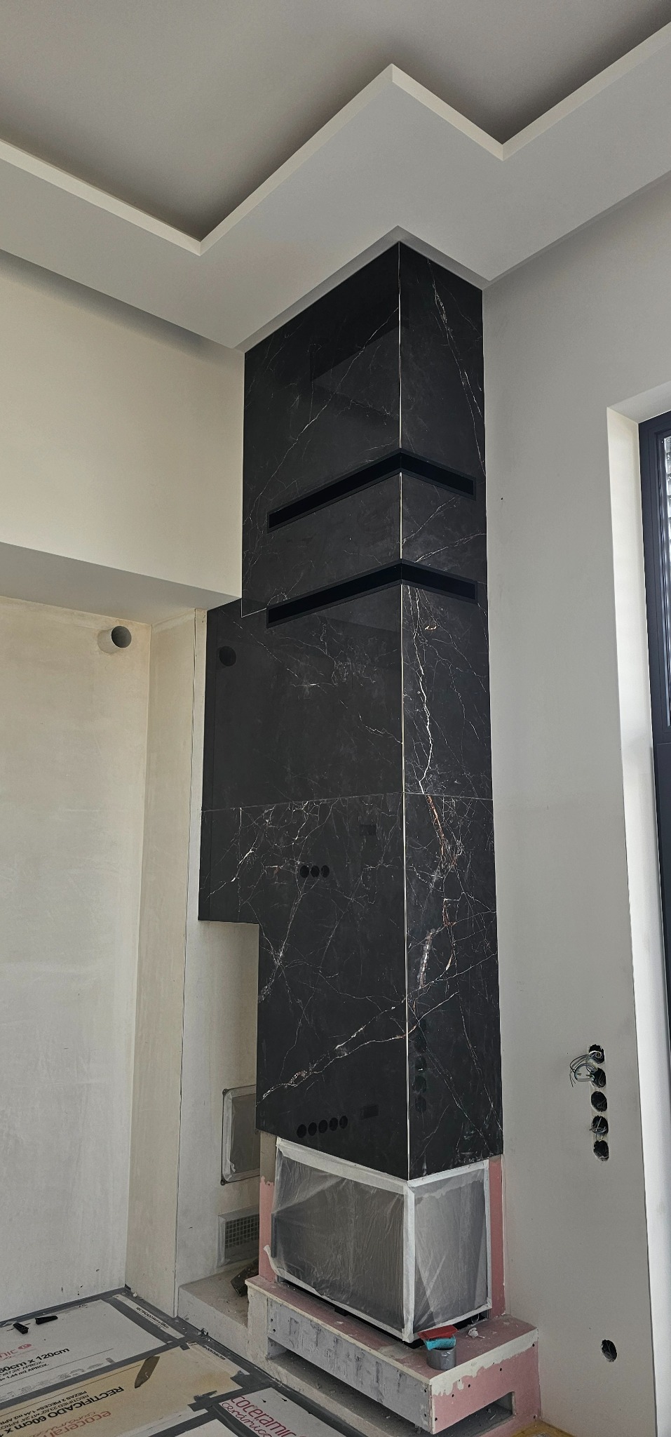 Wykończenie kominka czarnymi płytkami imitującymi marmur z widocznymi liniowymi kratkami wentylacyjnymi. Prace wykończeniowe w surowym wnętrzu.