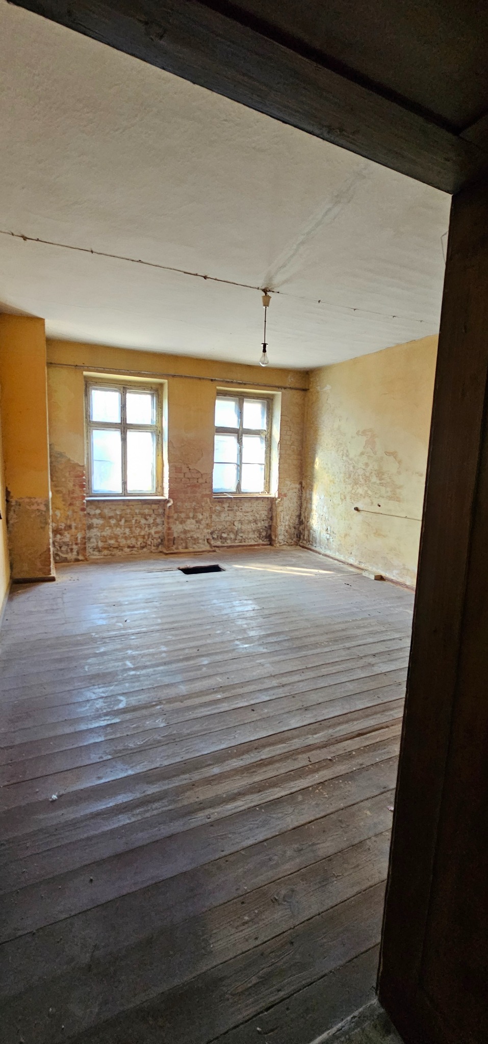 Wnętrze pokoju w trakcie remontu: odsłonięta cegła pod oknami, surowe drewniane deski podłogowe, żółte ściany z widocznymi śladami wcześniejszych warstw. Widok zza framugi.