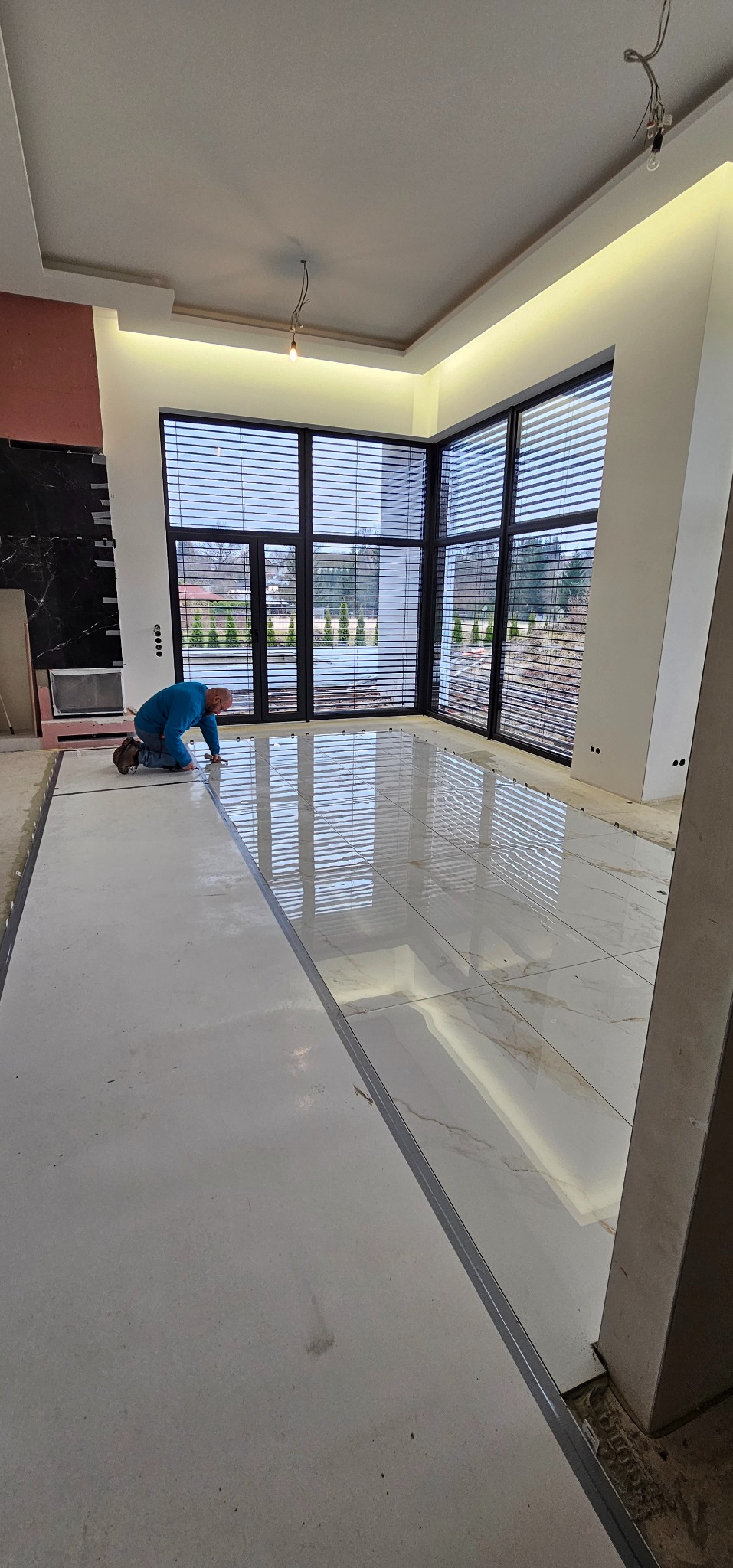 Glazurnik układa błyszczące, białe płytki podłogowe z delikatnym wzorem w przestronnym wnętrzu z dużymi oknami. Widoczne listwy przypodłogowe i narzędzia.