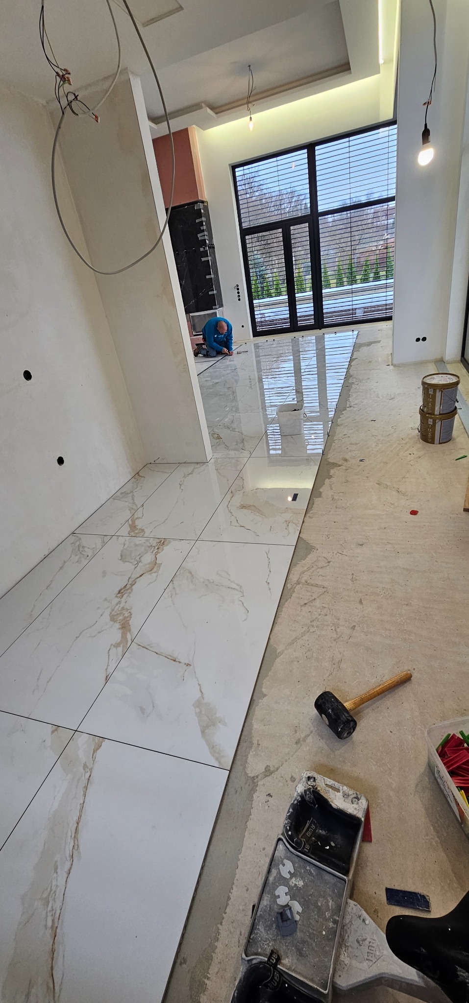 Układanie płytek podłogowych w przestronnym wnętrzu. Widoczne narzędzia glazurnicze i częściowo ułożone, białe płytki z marmurkowym wzorem. W tle okno i pracujący glazurnik.