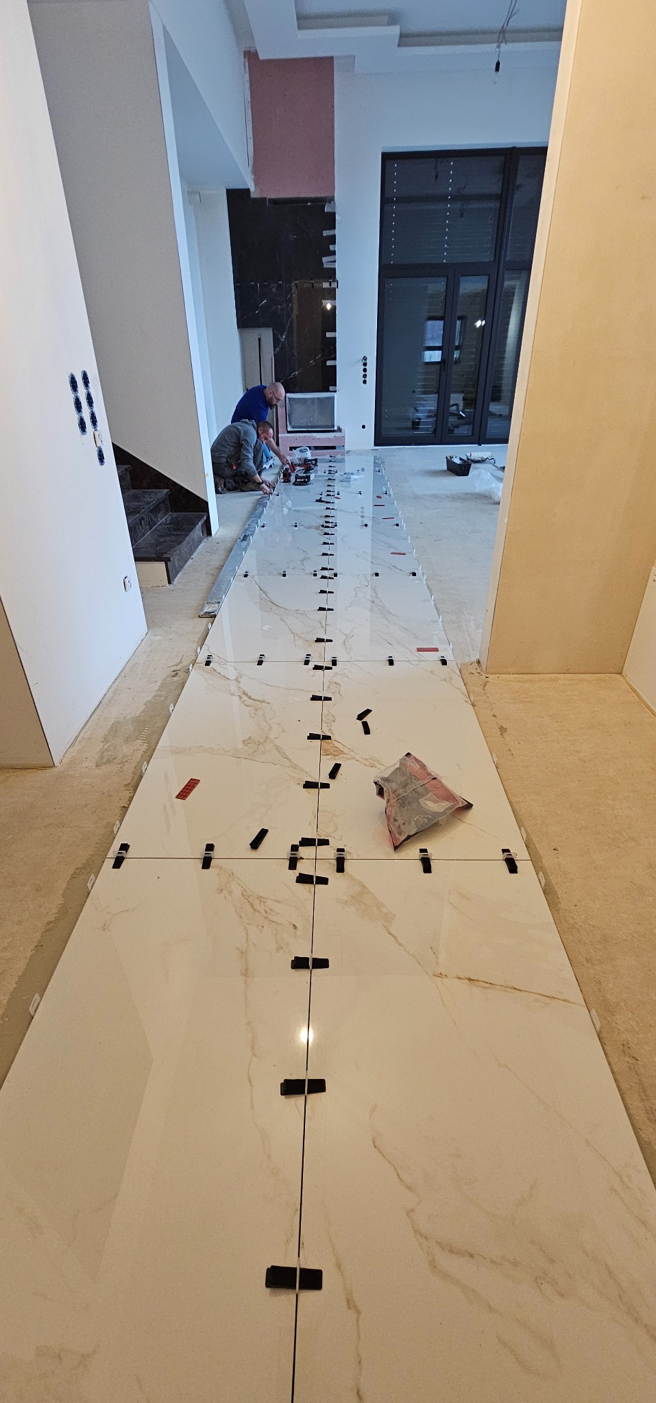 Układanie jasnych, marmurkowych płytek podłogowych z użyciem klipsów poziomujących. Dwóch glazurników pracuje w tle, widoczna część schodów i okna.