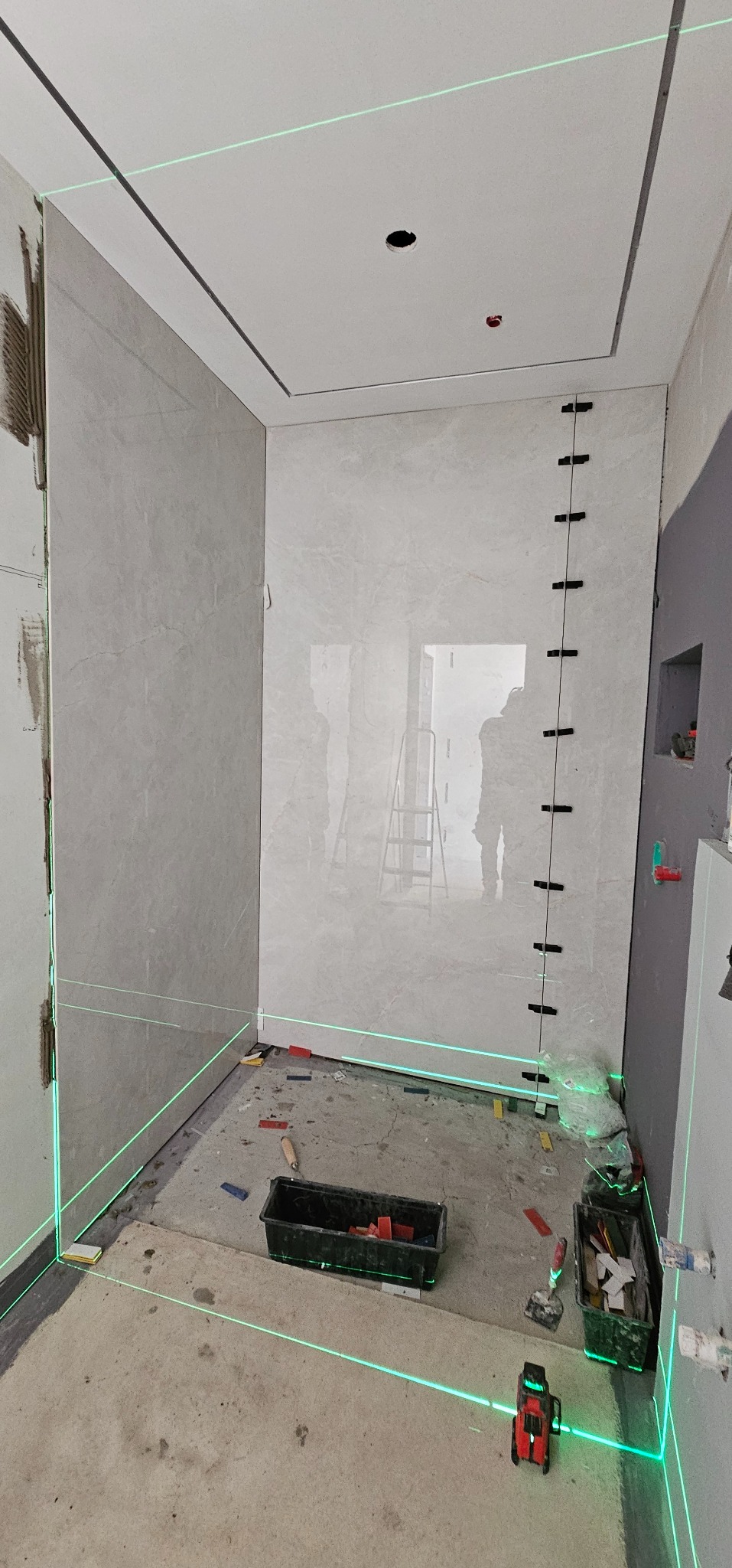 Łazienka w trakcie remontu: częściowo wyłożone płytki, poziomica laserowa wyznacza linie, widoczne narzędzia glazurnicze i akcesoria montażowe.