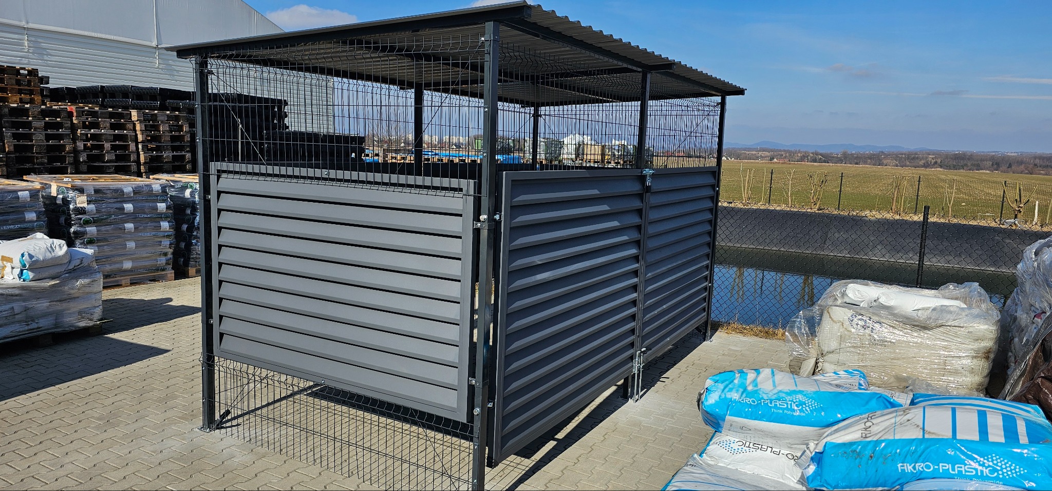 Szara, metalowa wiata śmietnikowa z ażurową górą i poziomymi lamelami, na kostce brukowej, w tle krajobraz. Widoczne worki z tworzywem.