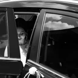 Panna młoda uśmiecha się, wyglądając z czarnego samochodu udekorowanego białą różą i wstążkami, widoczna przez otwarte drzwi.