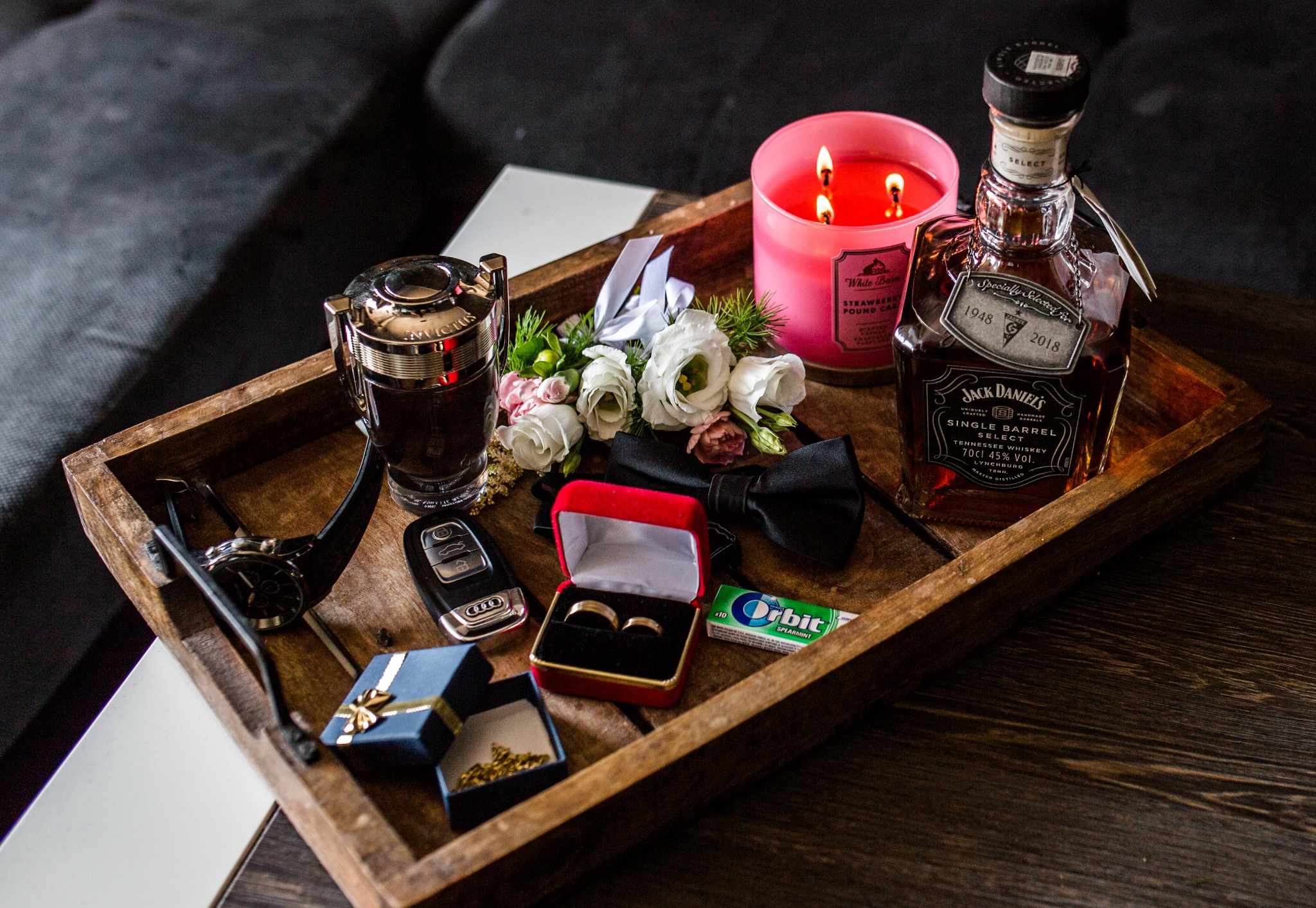 Przygotowania do ślubu: drewniana taca z obrączkami w czerwonym pudełku, bukiecikiem białych róż, muszką, butelką whisky Jack Daniels 1948-2018, świecą zapachową, perfumami Invictus, zegarkiem...