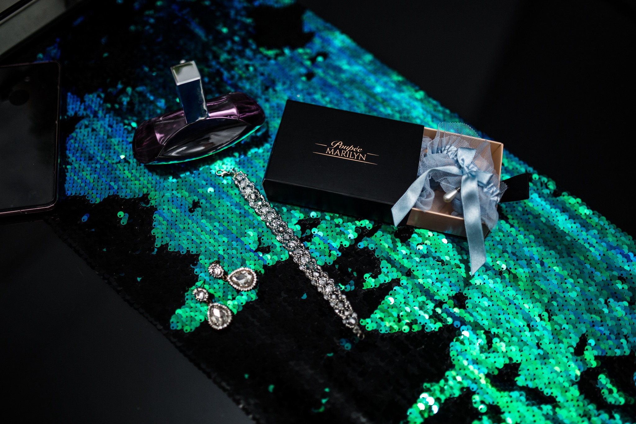 Kompozycja biżuterii ślubnej, w tym podwiązka w pudełku, kolczyki, bransoletka i flakon perfum, ułożona na błyszczącym, cekinowym tle w odcieniach turkusu i czerni, z widocznym fragmentem smartfona...