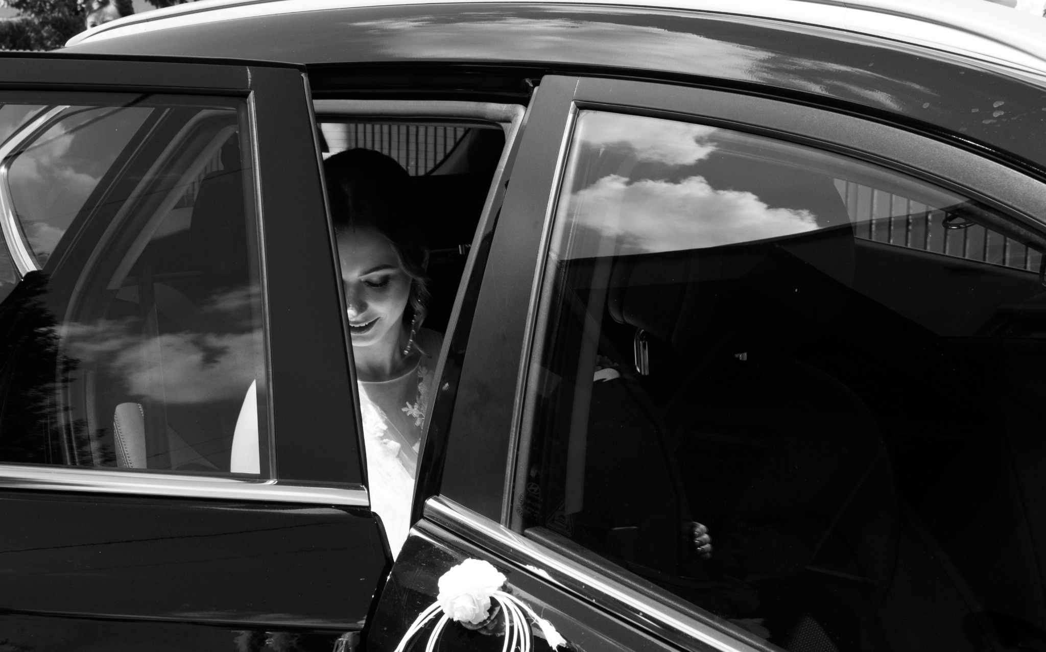 Panna młoda uśmiecha się, wyglądając z czarnego samochodu udekorowanego białą różą i wstążkami, widoczna przez otwarte drzwi.