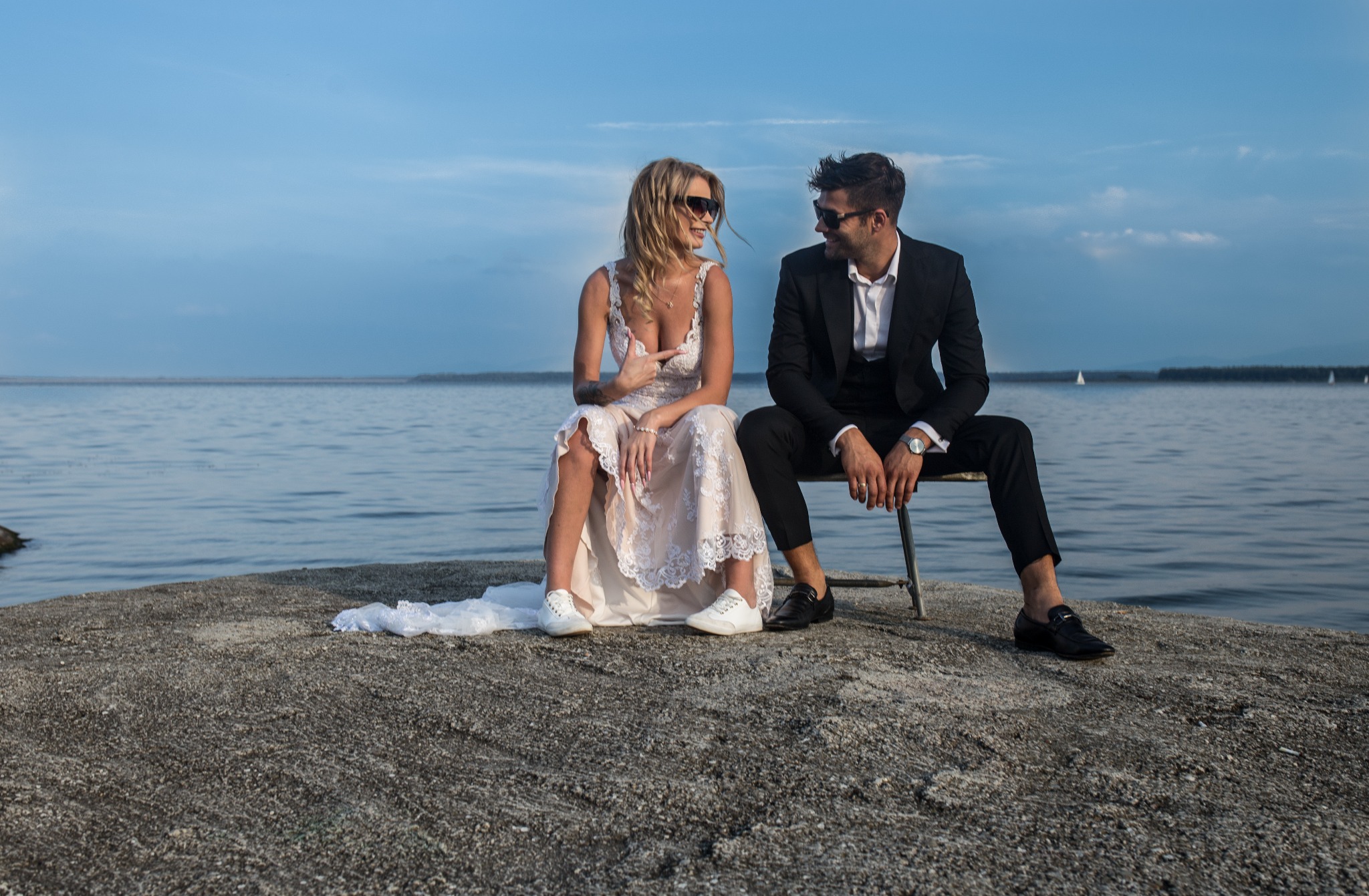 Para w strojach ślubnych siedzi na betonowym molo nad wodą, oboje w okularach przeciwsłonecznych, panna młoda w sukni z koronką i białych sneakersach, pan młody w czarnym garniturze i mokasynach,...