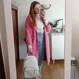 Kobieta robi selfie w lustrze, prezentując różowy płaszcz, biały sweter z warkoczami, dżinsy i biały plecak.