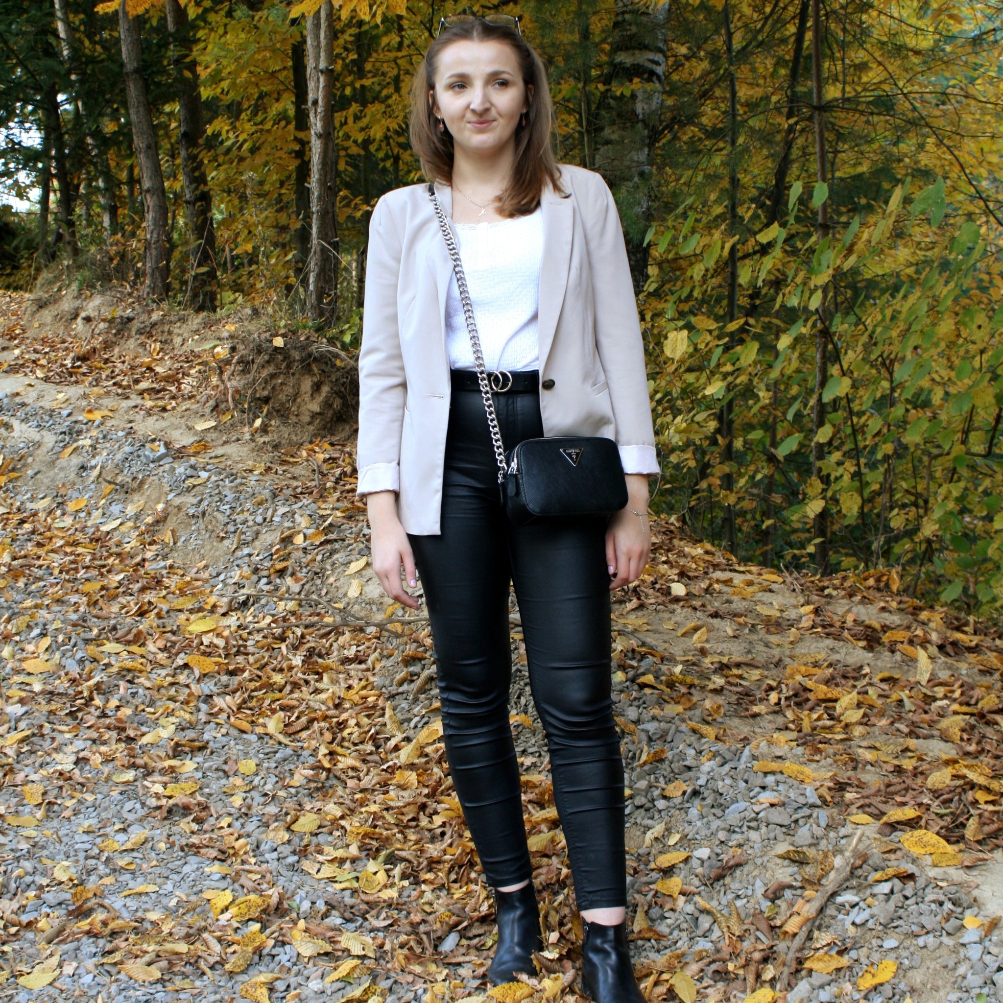 Portret kobiety w jesiennej scenerii, ubrana w beżową marynarkę, białą koszulkę, czarne skórzane spodnie i botki, z czarną torebką na łańcuszku, stojącej na ścieżce pokrytej liśćmi.