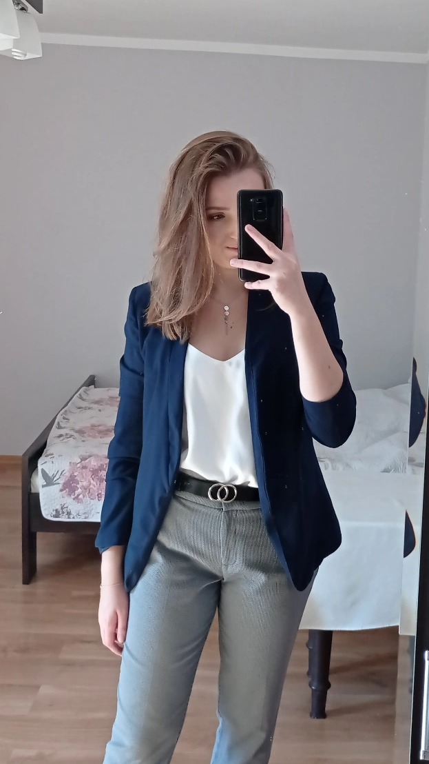 Młoda kobieta robi selfie w lustrze, ubrana w granatową marynarkę, białą bluzkę i szare spodnie z paskiem z podwójnym okręgiem. Lekko potargane blond włosy, subtelna biżuteria na szyi. W tle...