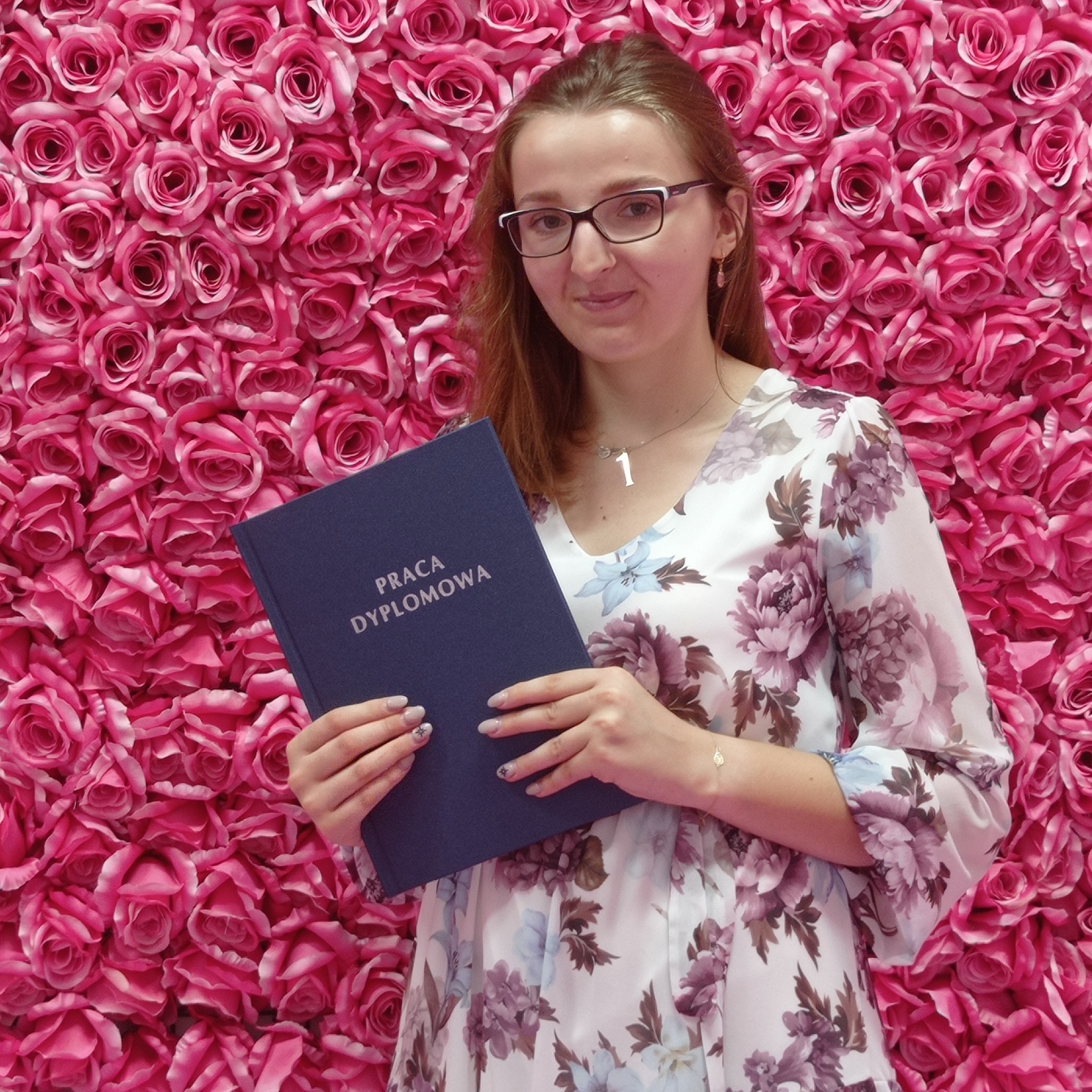 Młoda kobieta w okularach i kwiecistej sukience, trzymająca granatową oprawioną pracę dyplomową na tle ściany z różowych róż; delikatny manicure z motywem gwiazdek na paznokciach.