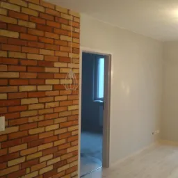 Ściana z cegły klinkierowej w nowym wnętrzu, z widocznym włącznikiem światła, lampą wiszącą i fragmentem podłogi laminowanej.