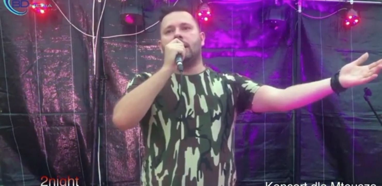 Wokalista w koszulce moro śpiewa do mikrofonu na scenie oświetlonej fioletowymi reflektorami, z tekstem 'Koncert dla Mteusza' widocznym w dolnym rogu.