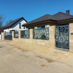 Nowoczesne ogrodzenie z metalową bramą o geometrycznym wzorze, osadzone na murku z jasnych bloczków, na tle dwóch domów i błękitnego nieba.