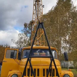 Żółta ciężarówka z wysoką, ażurową wieżą wiertniczą na tle jesiennych drzew i pochmurnego nieba.