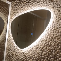 NeoPrąd - Owalne lustro z podświetleniem LED na ścianie wyłożonej kamieniami w łazience. Widoczna umywalka z miedzianą baterią i włącznik światła.