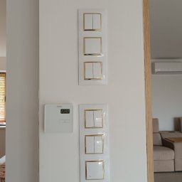 NeoPrąd - Biała ściana z zamontowanymi, nowoczesnymi włącznikami światła z złotymi ramkami i panelem sterowania ogrzewaniem podłogowym. Widok na wnętrze domu.