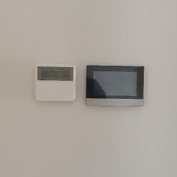 NeoPrąd - Na jasnej ścianie zamontowany panel sterowania ogrzewaniem i dotykowy panel domofonu w czarnej ramce. Minimalistyczny design, nowoczesne rozwiązania.