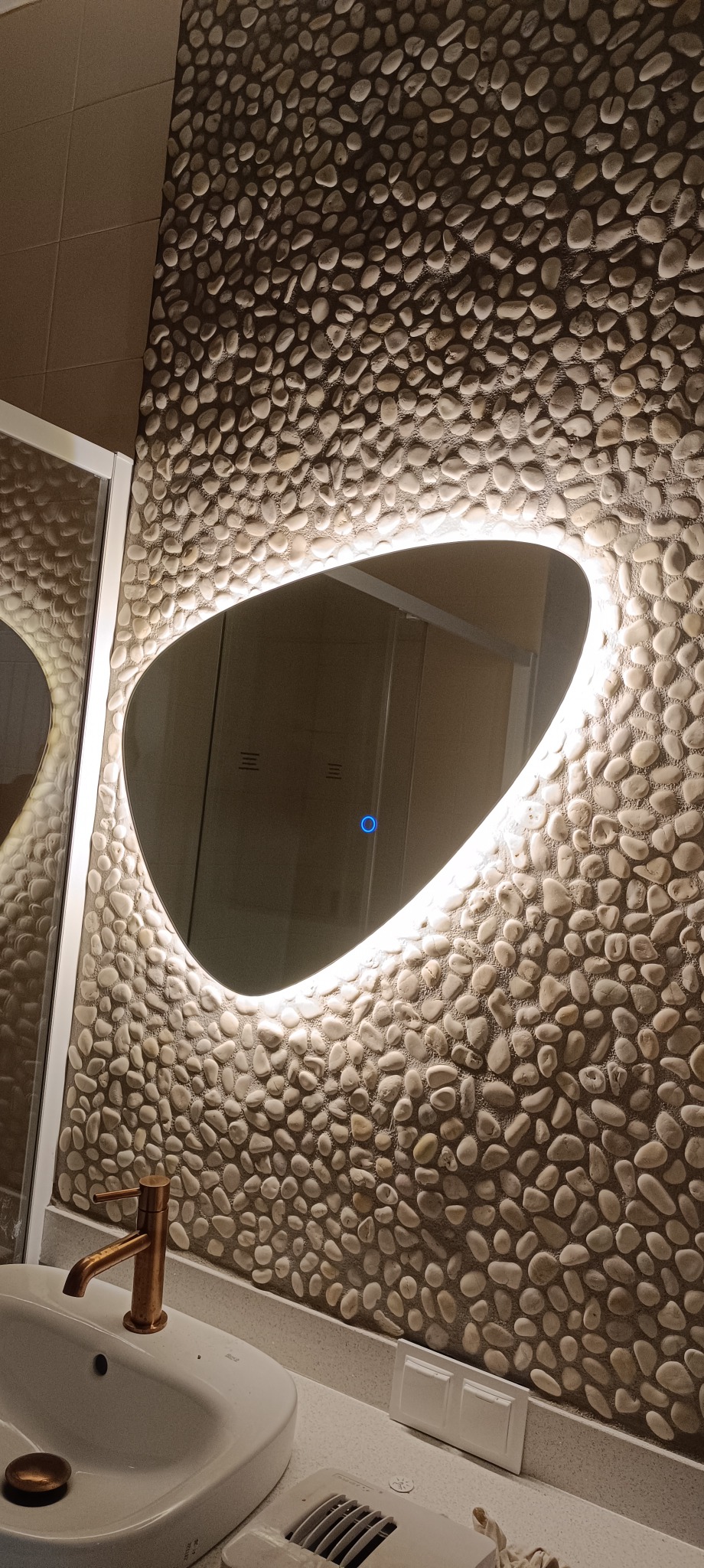 Owalne lustro z podświetleniem LED na ścianie wyłożonej kamieniami w łazience. Widoczna umywalka z miedzianą baterią i włącznik światła.