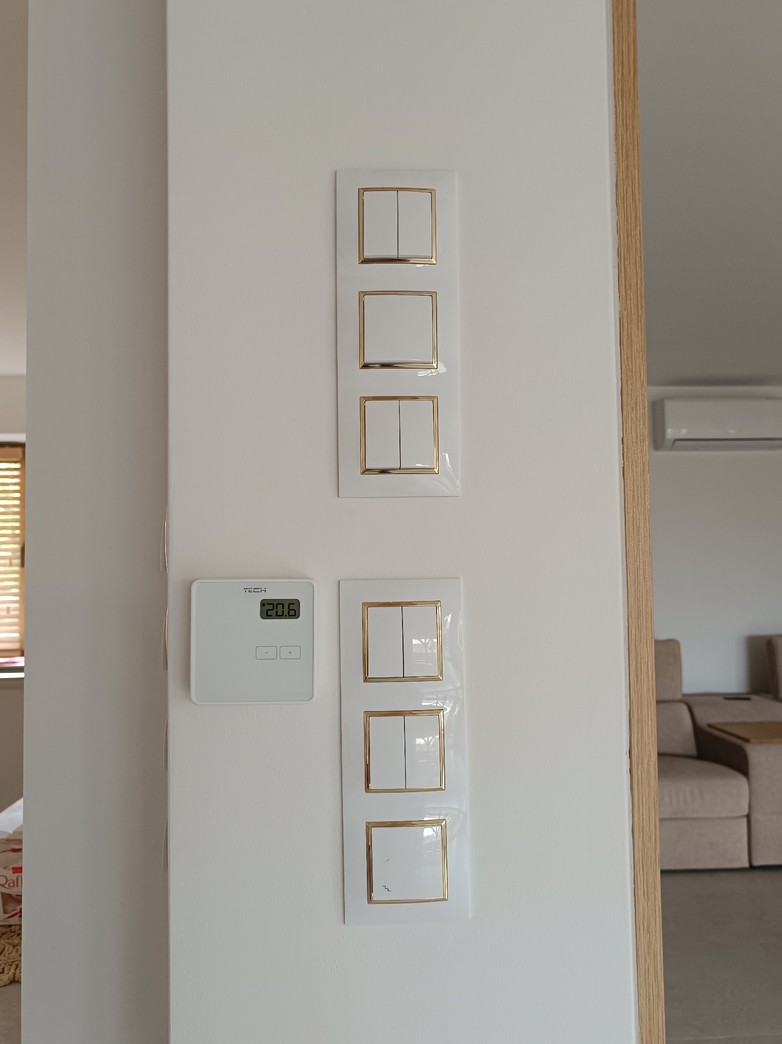 Biała ściana z zamontowanymi, nowoczesnymi włącznikami światła z złotymi ramkami i panelem sterowania ogrzewaniem podłogowym. Widok na wnętrze domu.