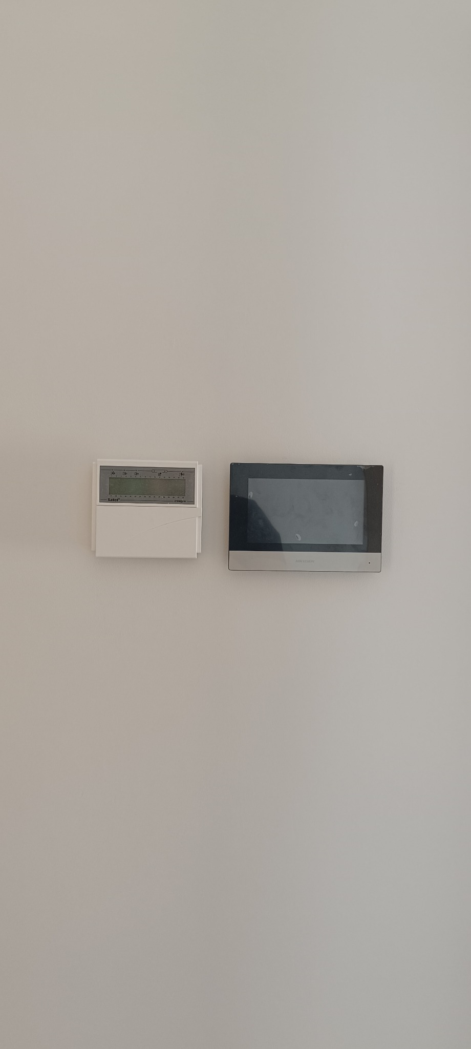 Na jasnej ścianie zamontowany panel sterowania ogrzewaniem i dotykowy panel domofonu w czarnej ramce. Minimalistyczny design, nowoczesne rozwiązania.