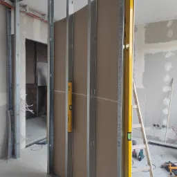 Montaż ściany działowej z płyty karton-gips na metalowym stelażu, widoczne profile, płyty G-K, poziomica na stelażu, drabina, narzędzia na podłodze w surowym wnętrzu.