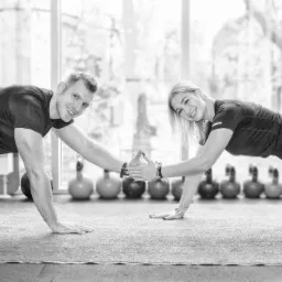 Czarno-białe zdjęcie przedstawia mężczyznę i kobietę wykonujących ćwiczenie deski w jasnym pomieszczeniu z rzędem odważników kettlebell w tle, dotykających dłońmi w geście współpracy.