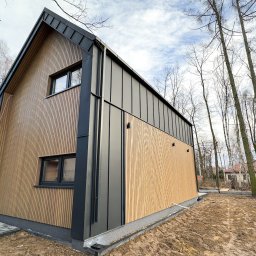 Usługi Stolarskie - Nowoczesny dom szkieletowy z elewacją z pionowych paneli drewnianych i grafitowymi elementami wykończeniowymi, widok z zewnątrz, otoczony drzewami.