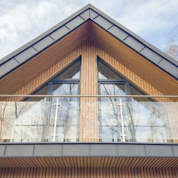 Usługi Stolarskie - Fragment elewacji domu z drewnianą fasadą, dwoma trójkątnymi oknami i balkonem z przezroczystą balustradą, widziany od dołu pod kątem.