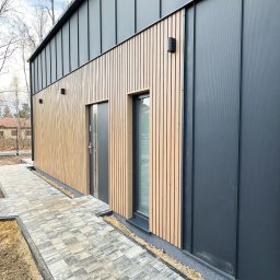 Usługi Stolarskie - Nowoczesny dom z elewacją z pionowych drewnianych paneli i grafitowych blach trapezowych, widok z boku na wejście i okno, z ułożoną kostką brukową wokół budynku.