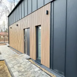 Nowoczesny dom z elewacją z pionowych drewnianych paneli i grafitowych blach trapezowych, widok z boku na wejście i okno, z ułożoną kostką brukową wokół budynku.