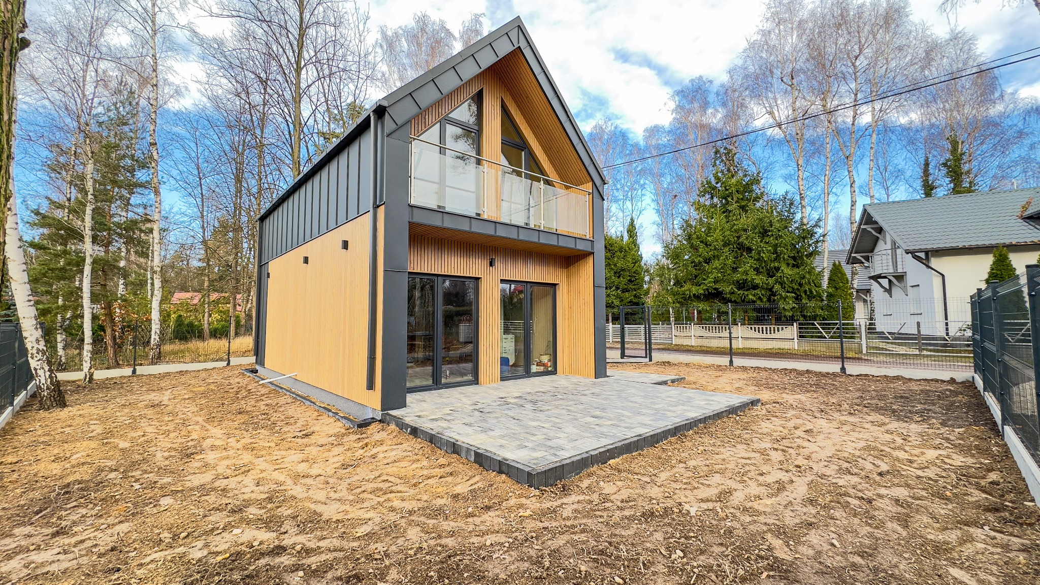 Nowoczesny, dwukondygnacyjny dom z drewnianą elewacją i grafitowym dachem, z balkonem z przeszkloną balustradą, na działce z częściowo ułożoną kostką brukową.