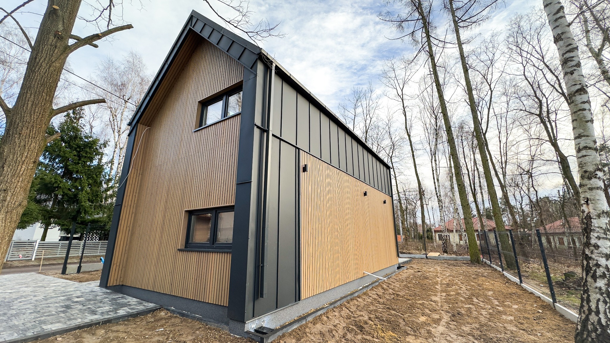 Nowoczesny dom szkieletowy z elewacją z pionowych paneli drewnianych i grafitowymi elementami wykończeniowymi, widok z zewnątrz, otoczony drzewami.