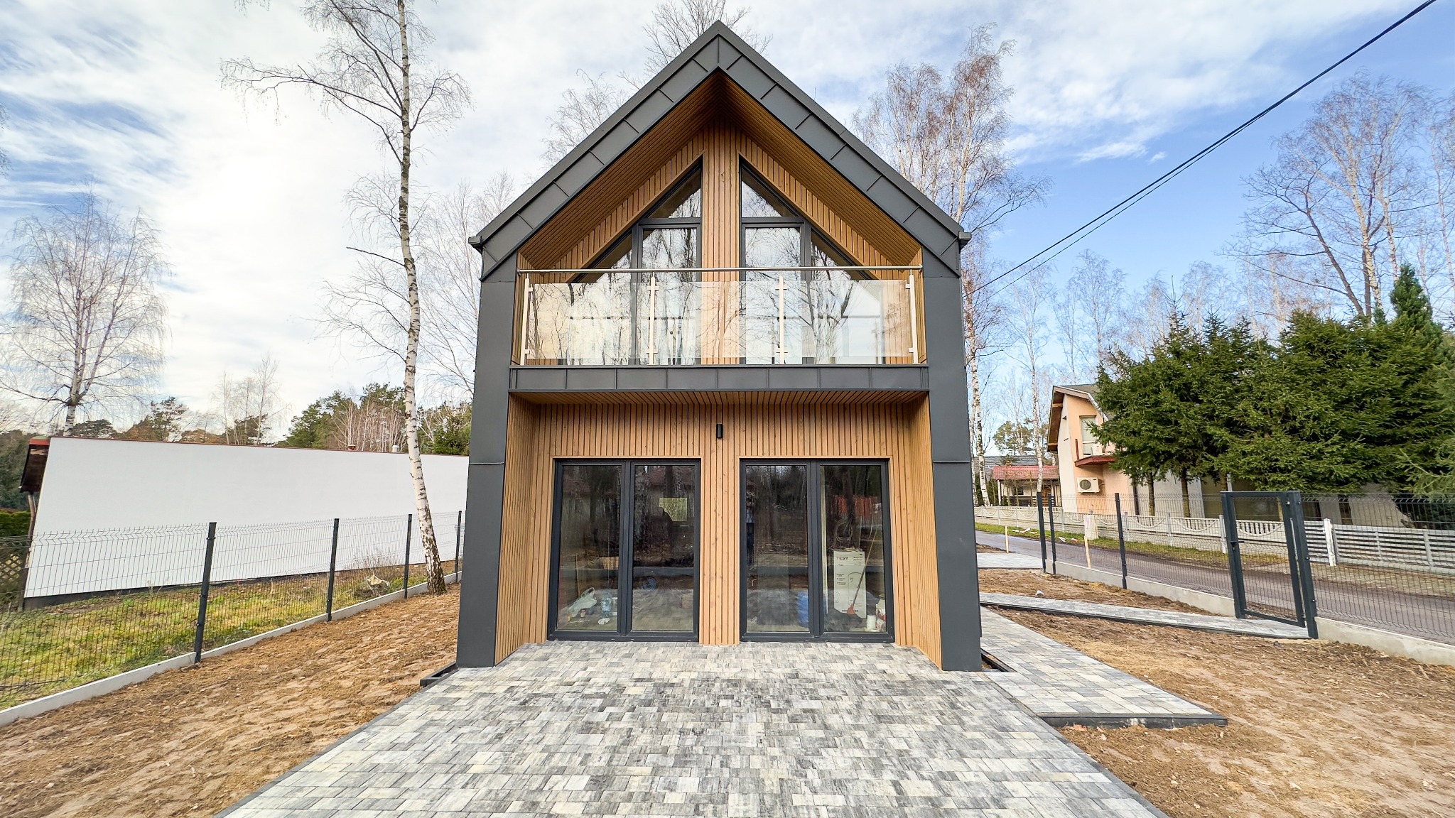 Dwukondygnacyjny dom z drewnianą elewacją i balkonem z przezroczystą balustradą, widok z przodu na nowo ułożony chodnik z kostki brukowej.