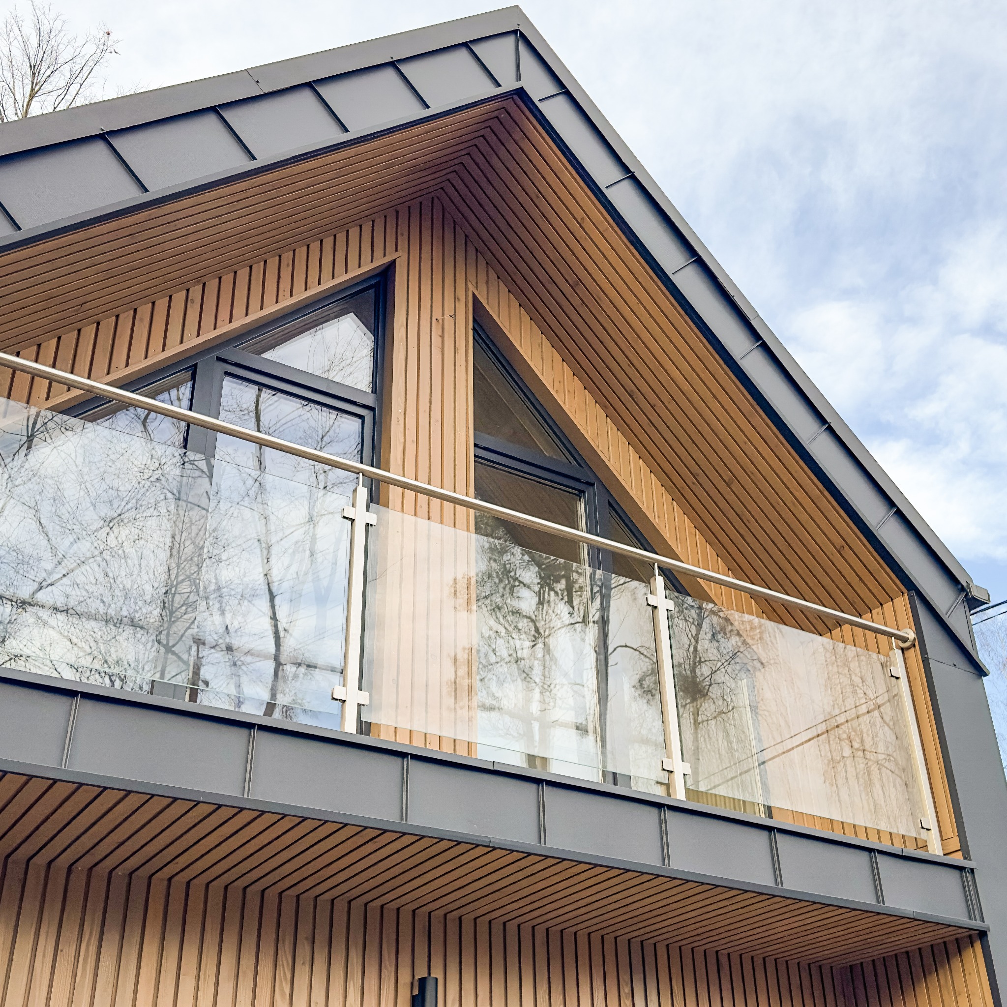 Nowoczesny dom z drewnianą elewacją i szarym dachem, z balkonem z przezroczystą szklaną balustradą i metalowymi poręczami, odbicie drzew w oknach.