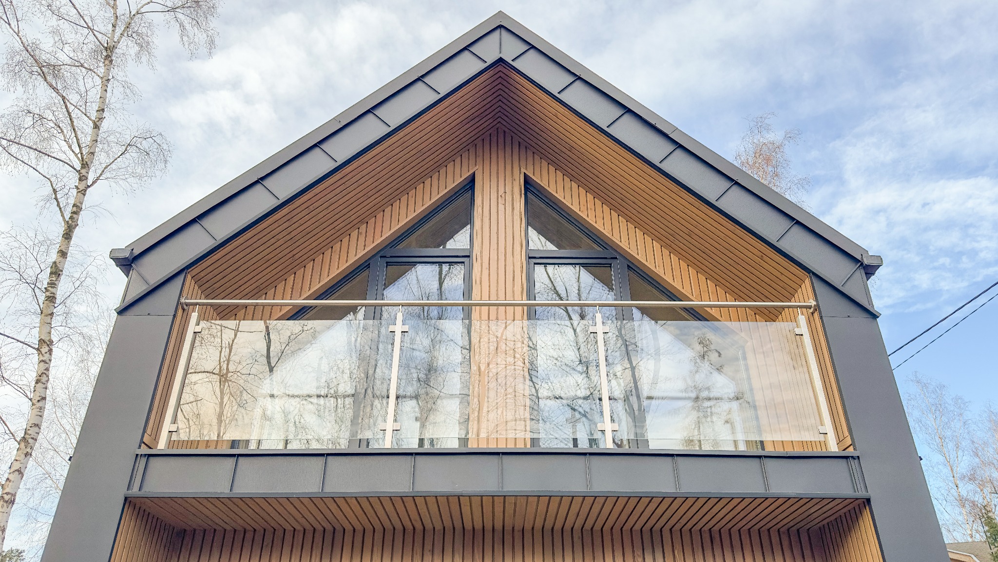 Fragment elewacji domu z drewnianą fasadą, dwoma trójkątnymi oknami i balkonem z przezroczystą balustradą, widziany od dołu pod kątem.