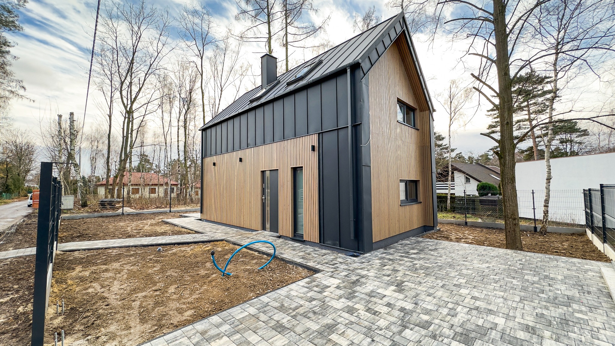 Nowoczesny dom z drewnianą elewacją i ciemnym metalowym dachem, widok z zewnątrz na nowo wybrukowany podjazd i częściowo zagospodarowany teren.
