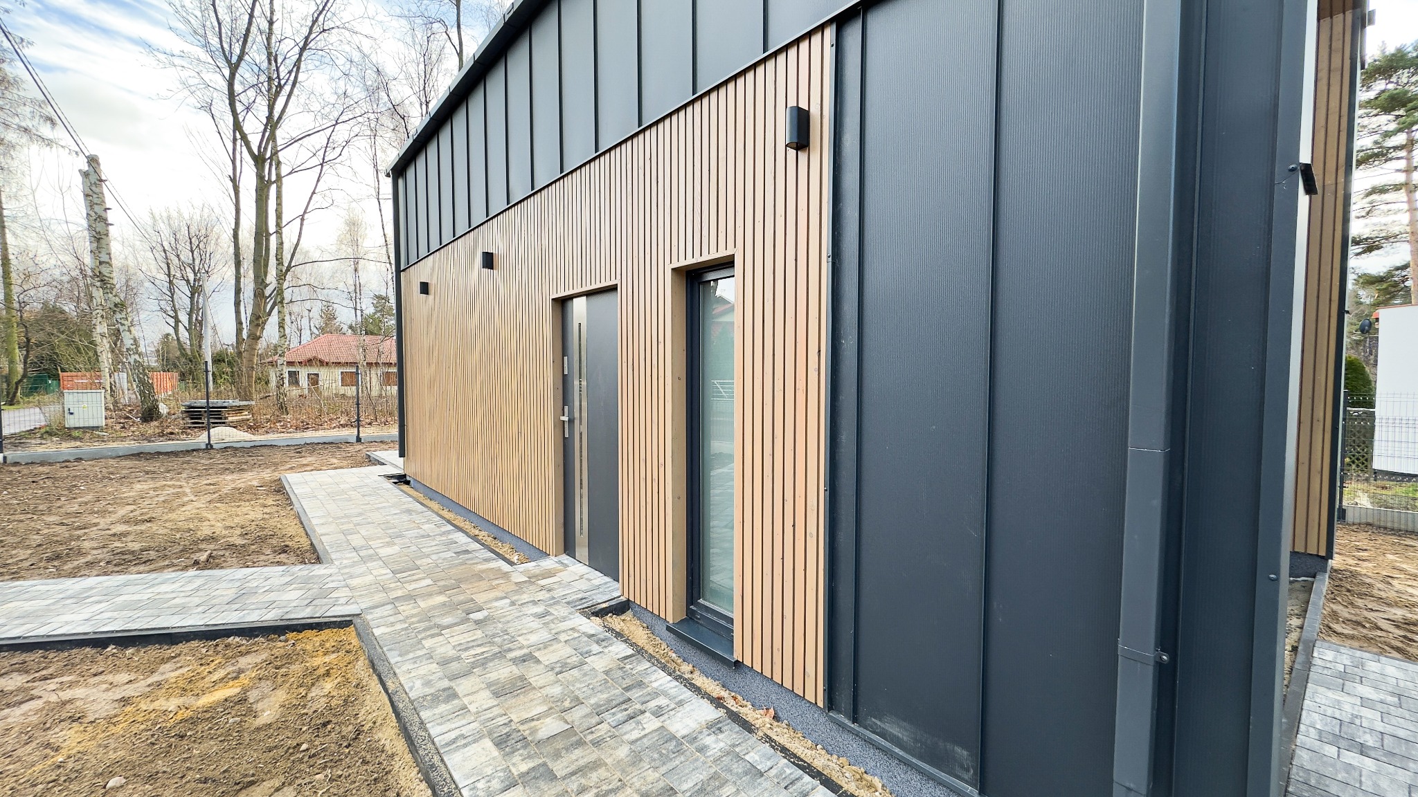 Nowoczesny dom z elewacją z pionowych drewnianych paneli i grafitowych blach trapezowych, widok z boku na wejście i okno, z ułożoną kostką brukową wokół budynku.