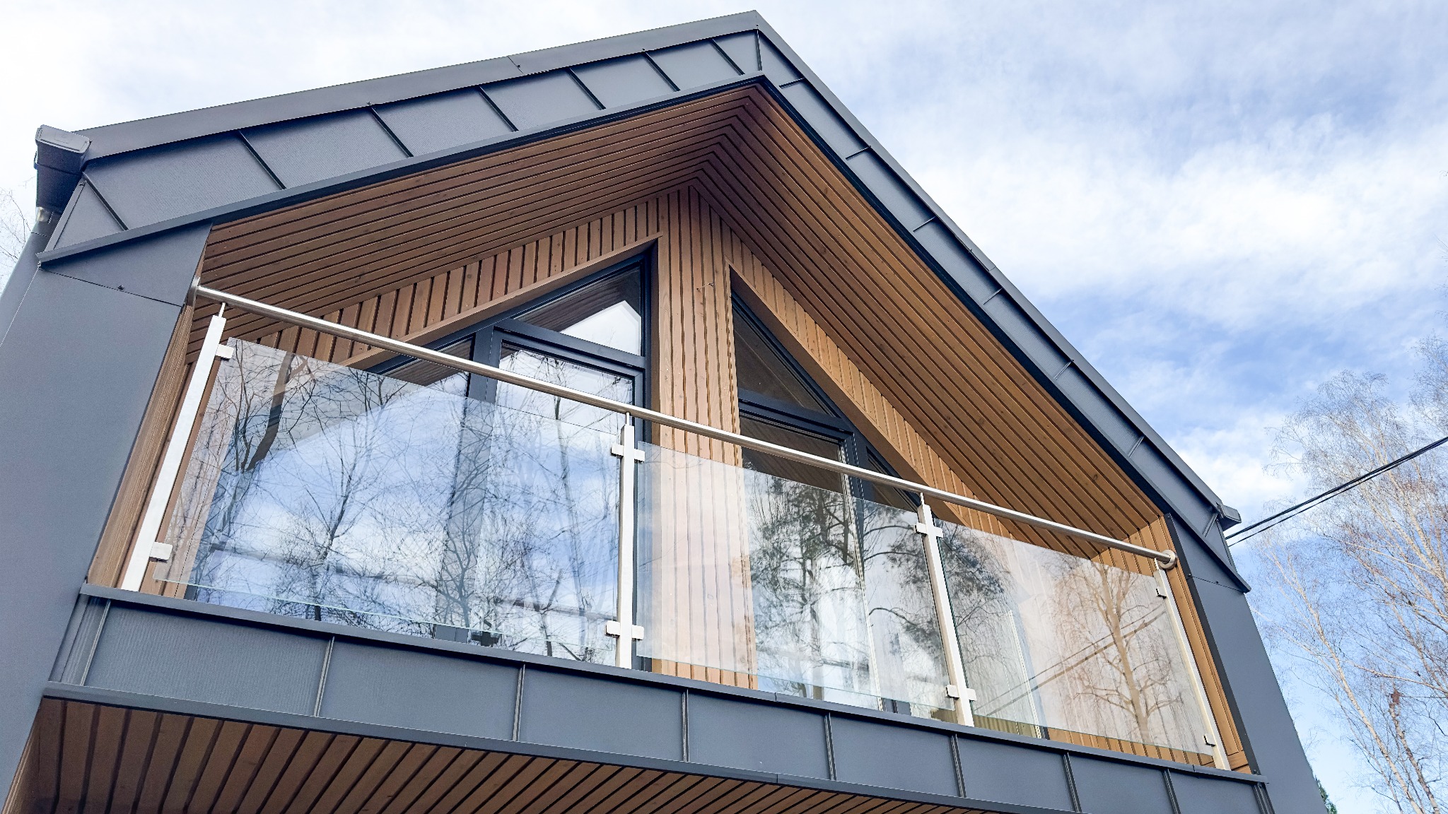 Nowoczesny dom z balkonem o szklanej balustradzie i drewnianej elewacji, widok z dołu na błękitne niebo.