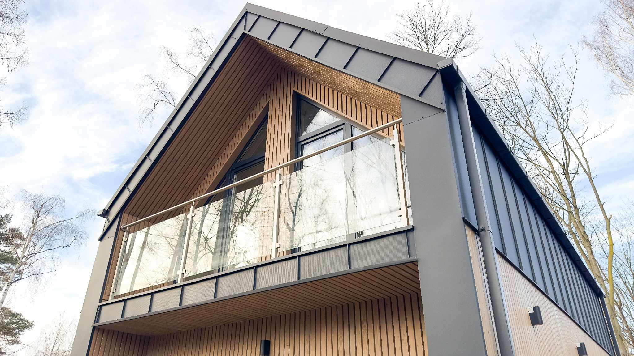 Nowoczesny dom z drewnianą elewacją i szklaną balustradą balkonu, uchwycony pod kątem z dołu, z widocznymi drzewami w tle.