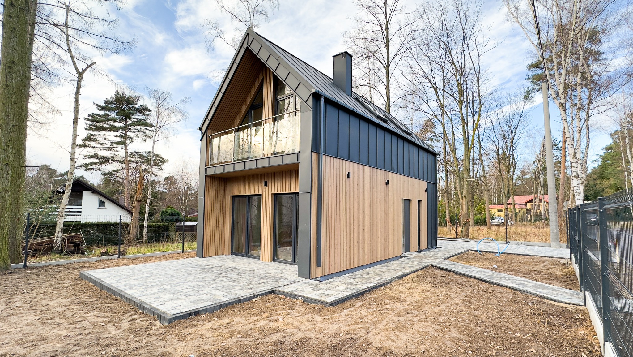 Nowoczesny dom z dwuspadowym dachem i balkonem ze szklaną balustradą, wykończony drewnianą elewacją i ciemną blachą, otoczony drzewami i świeżo ułożoną kostką brukową.
