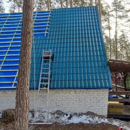 May bud - Dach domu w trakcie krycia: połowa z niebieską dachówką, druga z folią i łatami. Drabina oparta o ścianę. W tle las i fragment osoby.