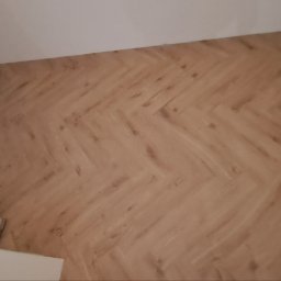 May bud - Ściana wyłożona panelami w jodełkę, widoczne kartony po panelach przy podłodze. Białe ściany i sufit. Ujęcie z dołu, lekko z ukosa.